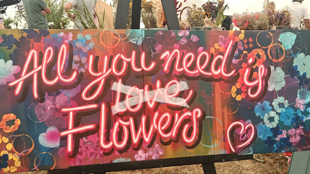 A colourful sign of painted flowers with the words 'all you need is flowers' written in not-neon pink lettering