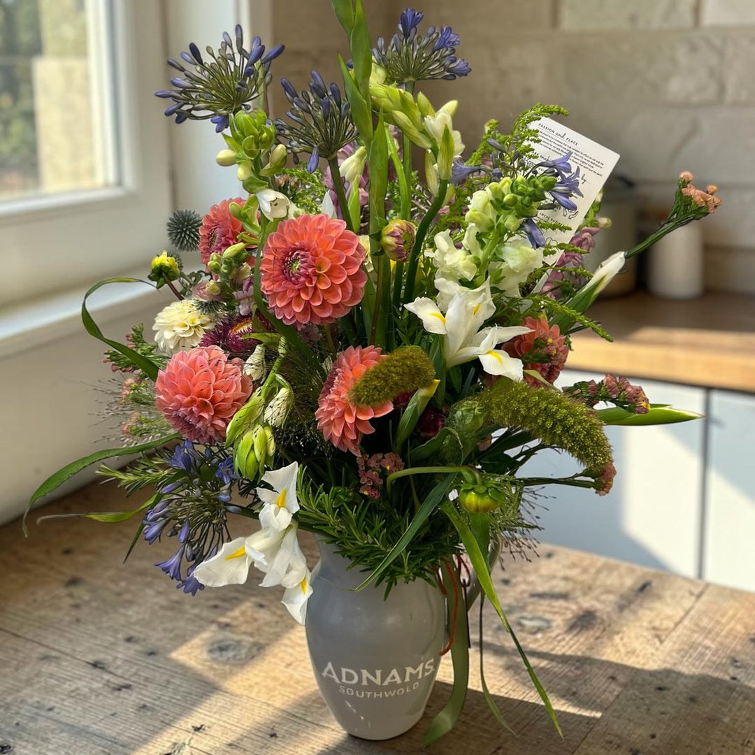 A vibrant wildflower arrangement in a gray ceramic vase printed with 'ADNAME' and 'Florist', set on a rustic wooden table in a sunlit room with a stone wall background.