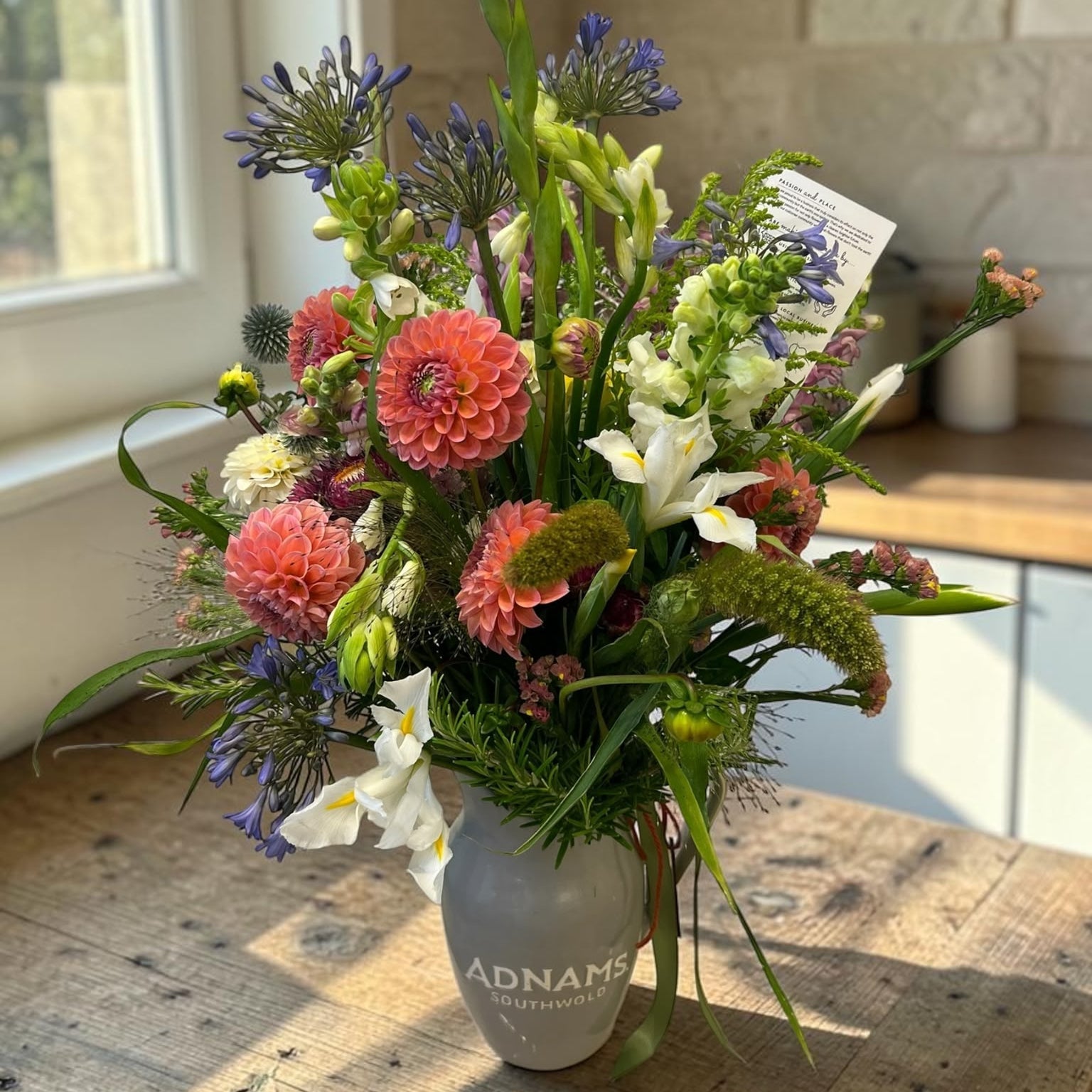 A vibrant wildflower arrangement in a gray ceramic vase printed with 'ADNAME' and 'Florist', set on a rustic wooden table in a sunlit room with a stone wall background.