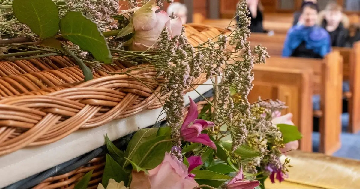 A close-up of a woven wicker basket lined with white material and decorated with green leaves, dried flowers, and pink blossoms, with a softly blurred church interior and seated people in the background.