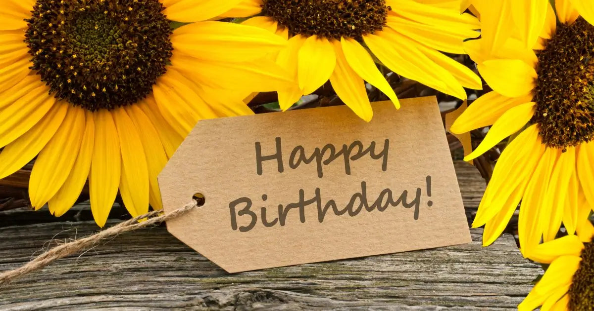 A brown kraft paper birthday tag with 'Happy Birthday!' written in black script, tied with twine and placed among bright yellow sunflowers on a rustic wooden surface.
