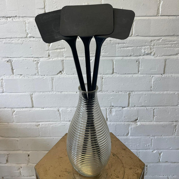 A black plastic angled head label placed on a metal vase, set against a white brick background.