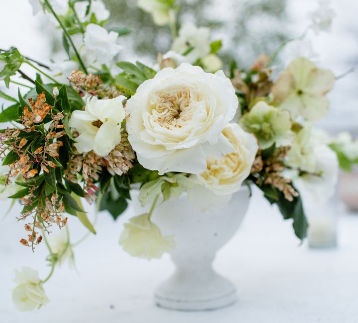 A white floral arrangement featuring large cream and white roses with green foliage and small dried orange-brown accents, presented in a white pedestal vase.