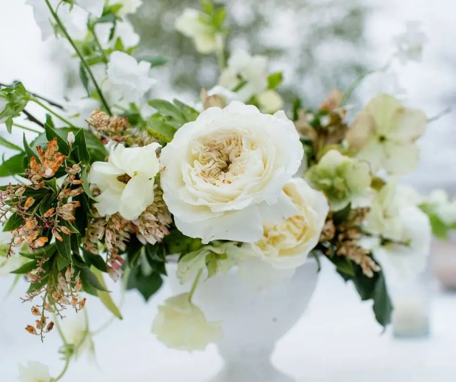 A close-up of a white floral arrangement featuring cream-colored roses, white blossoms, and green foliage in a white vase against a soft, blurred light background.