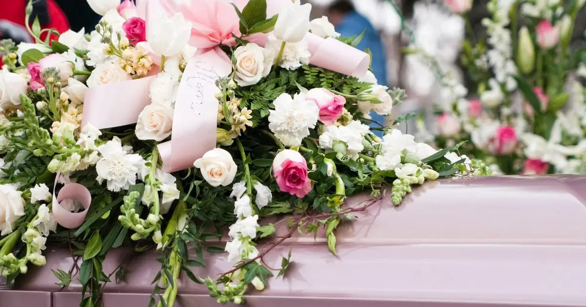 A floral arrangement with white and pink flowers, green foliage, and pale pink ribbons draped over a light pink casket in an outdoor setting.