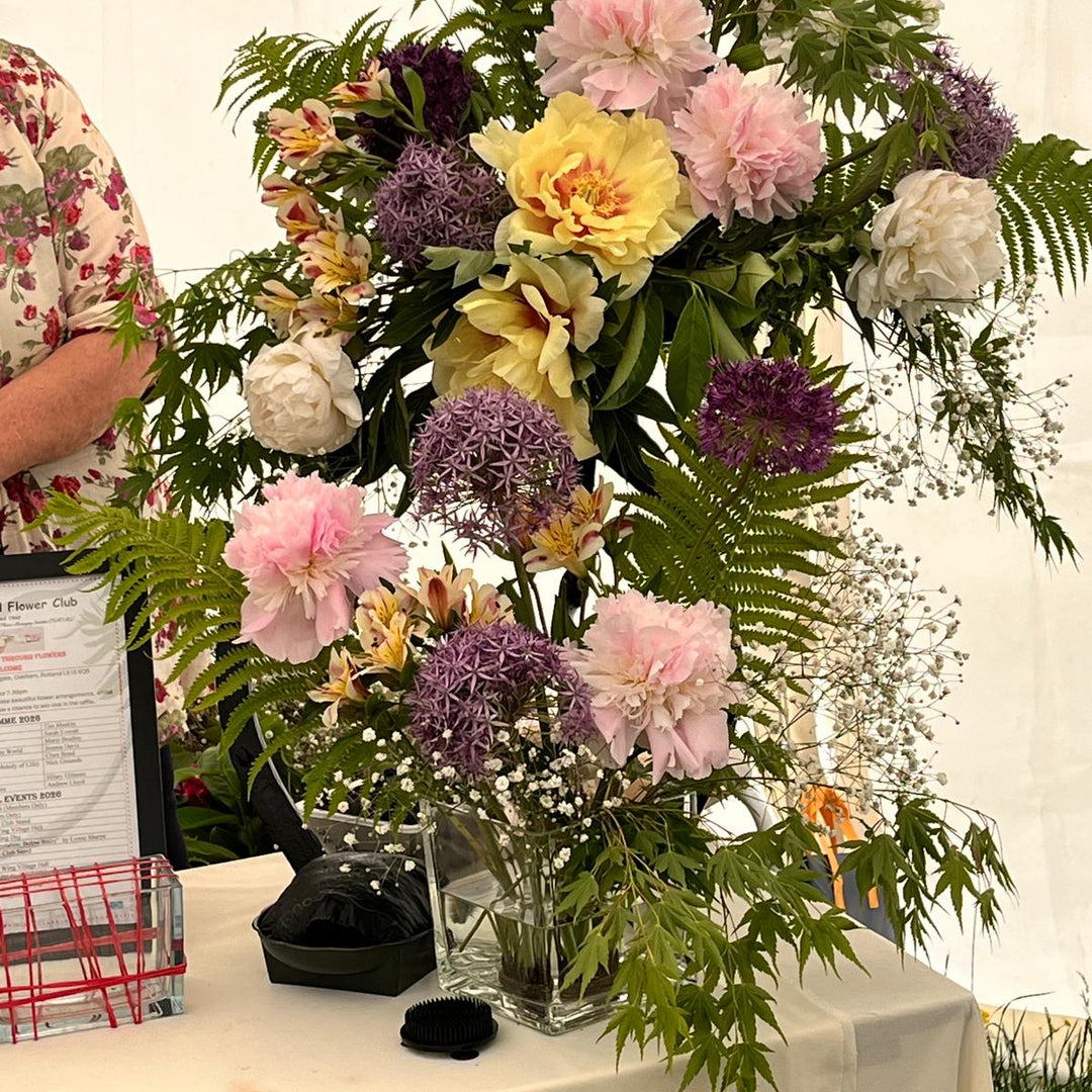 A large floral arrangement with pink, yellow, and purple flowers and green foliage displayed on a white tablecloth-covered table. Part of a person in a floral shirt is visible at the left edge, at the Rutland Flower Club Show