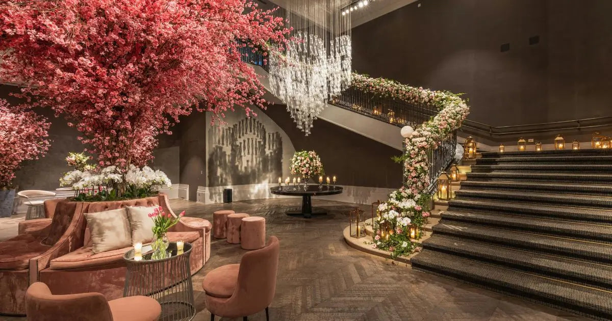 A luxurious indoor corporate space featuring a grand staircase decorated with white flowers, a large pink cherry blossom tree, velvet lounge seating, and a crystal chandelier.