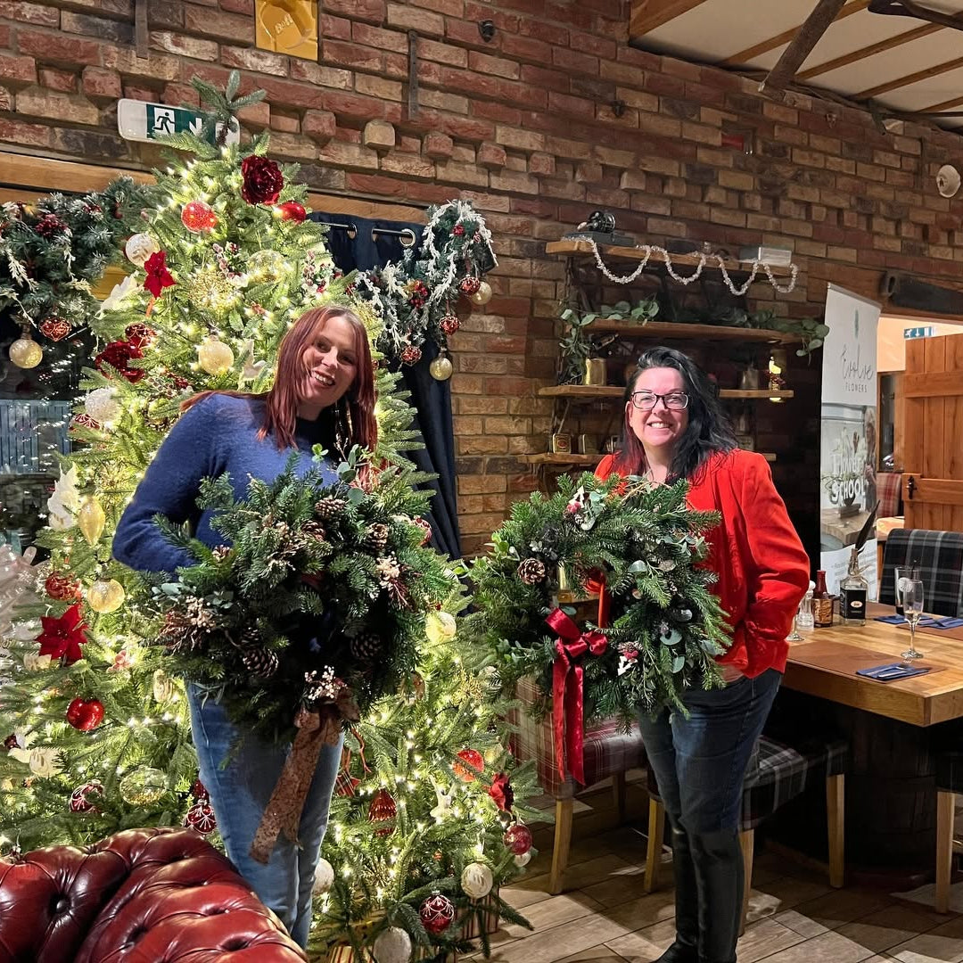 Two attendees posing with handmade Christmas door wreaths in front of a festive Christmas tree and brick fireplace.