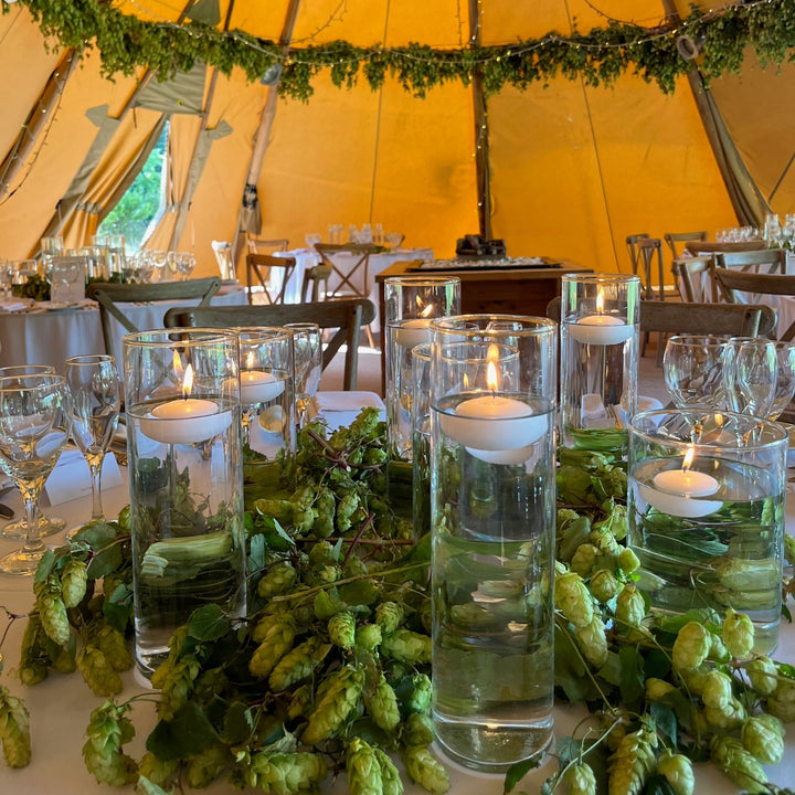 The warm interior of a large event tipi set up for a wedding reception. Floating candles in tall glass cylinders illuminate the foreground table, surrounded by a rustic runner of fresh, vibrant green hop flowers and leaves. Garlands of matching fresh hops and warm fairy lights are draped overhead across the canvas and wood frame structure.