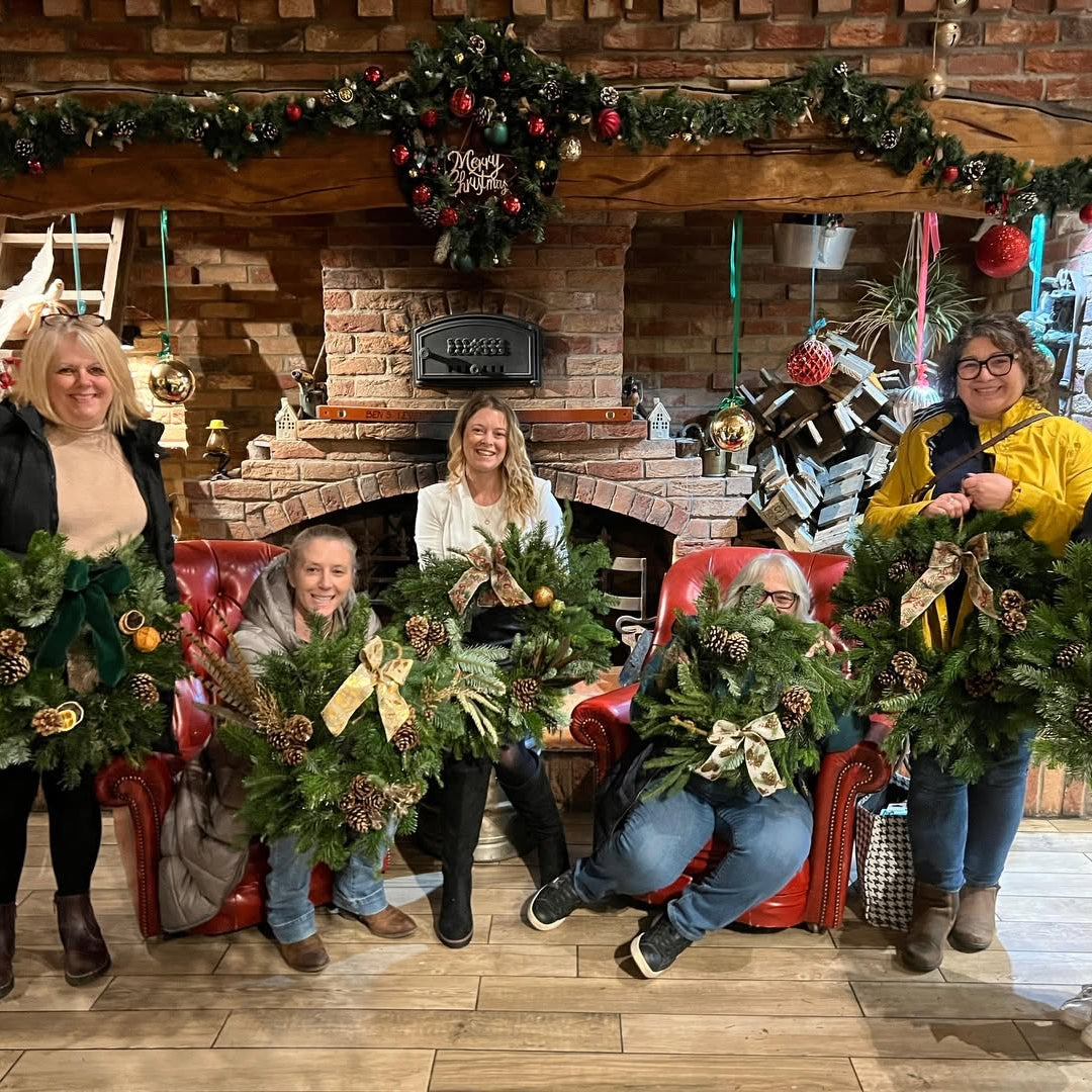 Five women sitting on red leather armchairs holding finished Christmas wreaths decorated with red velvet bows and dried elements.