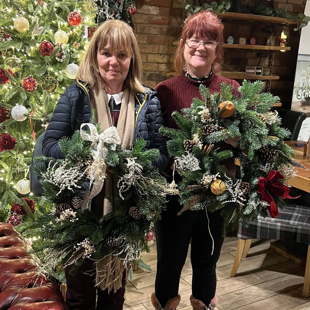 Two attendees posing with handmade Christmas door wreaths in front of a festive Christmas tree and brick fireplace.