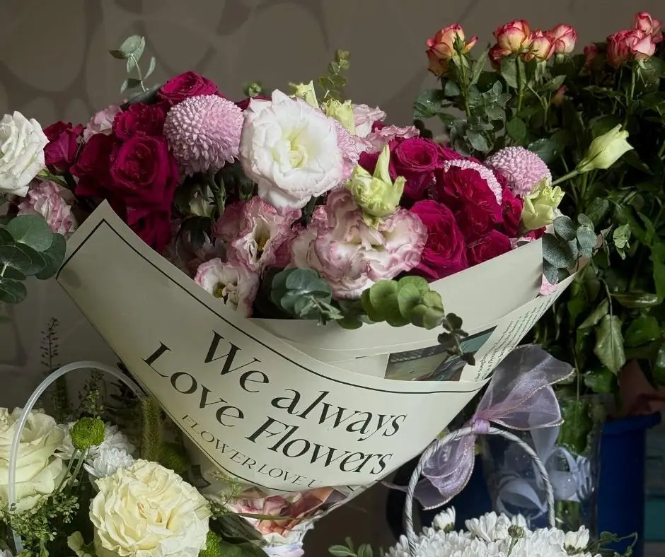 A close-up of a flower bouquet wrapped in cream paper printed with the words 'We always Love Flowers'. The bouquet from Evolve Flowers features deep pink, magenta, and white flowers, including roses, chrysanthemums, and lisianthus, with green foliage and eucalyptus stems.