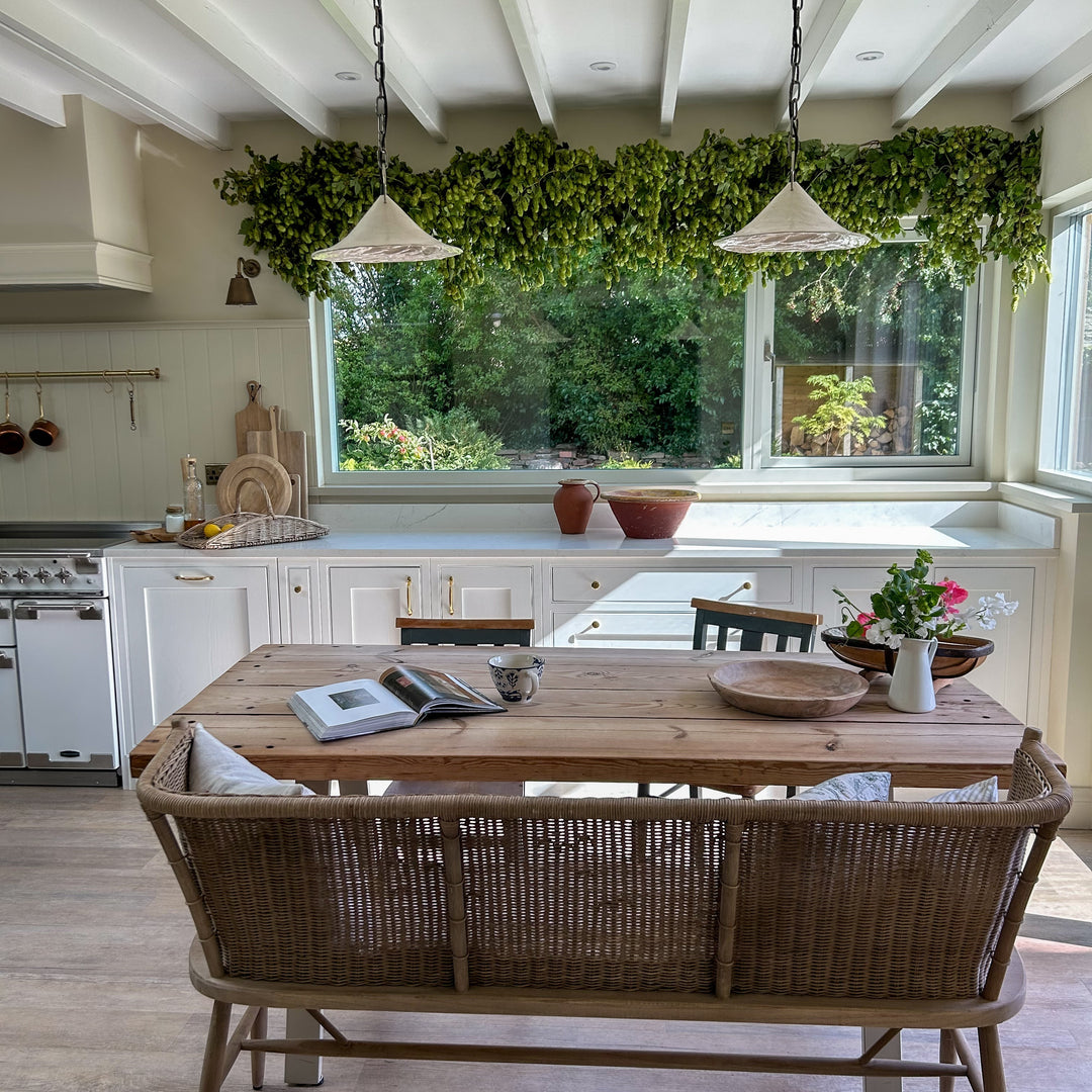 A rustic white kitchen featuring a large overhead garland of dense green fresh hop bines draped across a wide window frame above a wooden dining table.