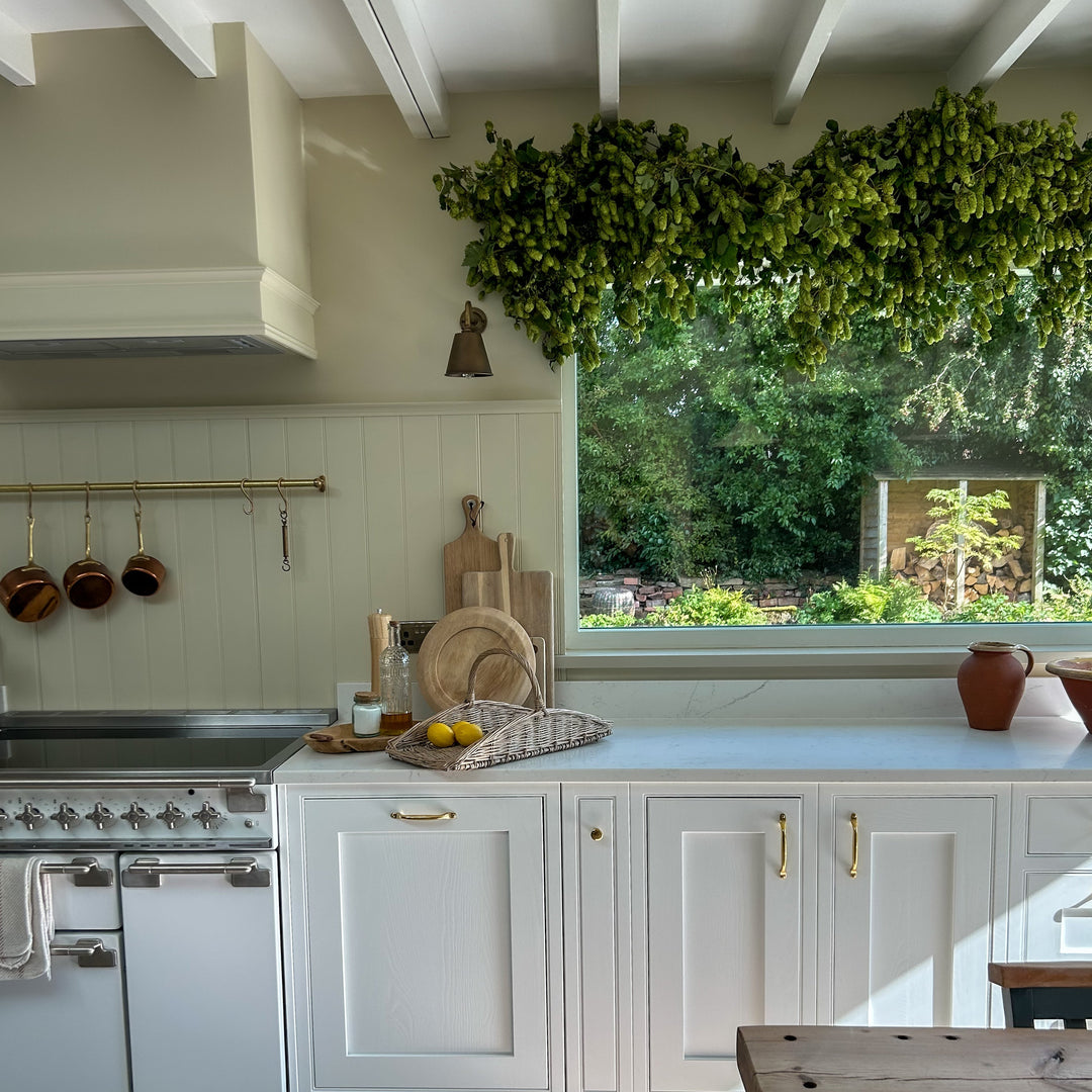 A bright, modern kitchen interior with white shaker-style cabinets, a white range stove, a large window looking out to a garden, and a wooden dining table in the foreground.