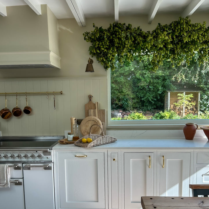 A bright, modern kitchen interior with white shaker-style cabinets, a white range stove, a large window looking out to a garden, and a wooden dining table in the foreground.
