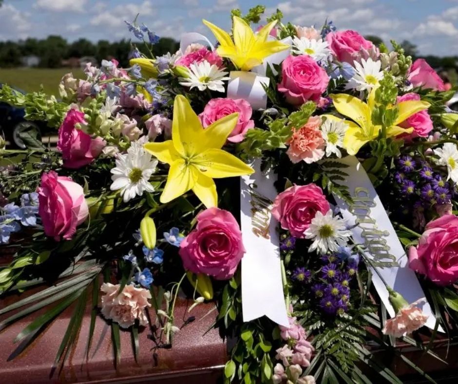 A vibrant floral arrangement for a casket, featuring pink roses, yellow lilies, white daisies, and blue flowers with white memorial ribbons, displayed outdoors on a casket.