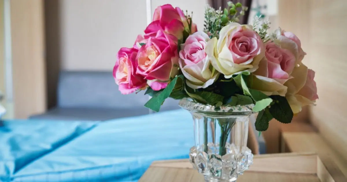 A bouquet of pink and cream roses with green leaves in a clear glass vase, placed on a wooden table in a softly blurred hospital room setting with a blue bedspread in the background.