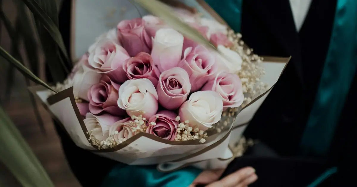 A close-up of a bouquet of pink and white roses wrapped in white paper with gold trim, held by a person wearing a graduation gown.