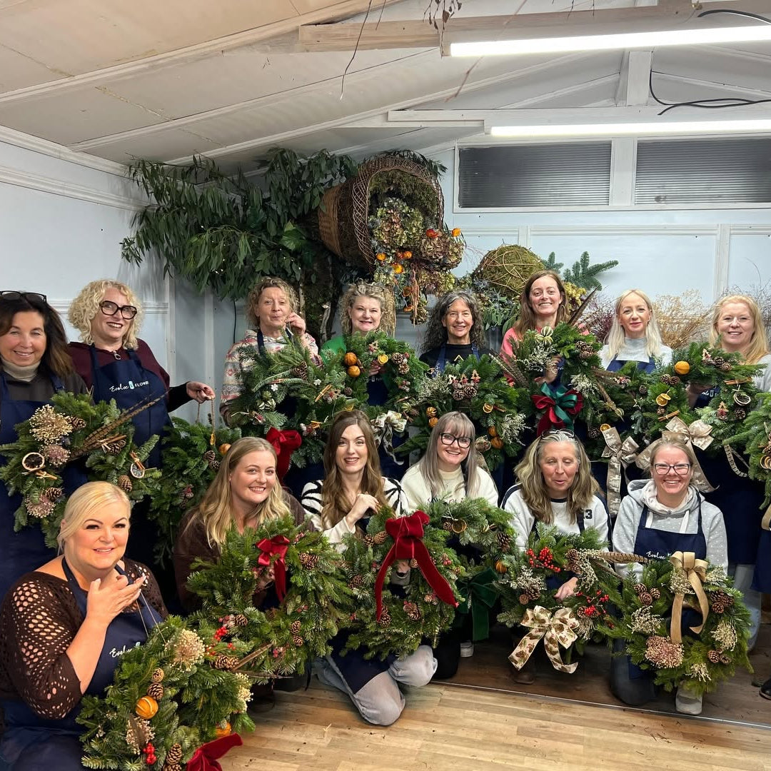 A group of happy participants posing with their handmade festive Christmas wreaths inside a floral workshop studio.