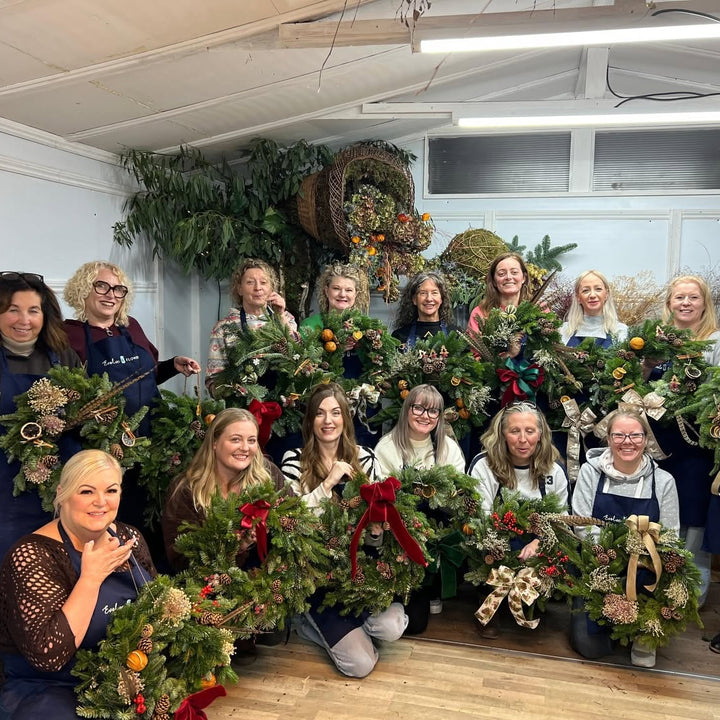 A group of happy participants posing with their handmade festive Christmas wreaths inside a floral workshop studio.