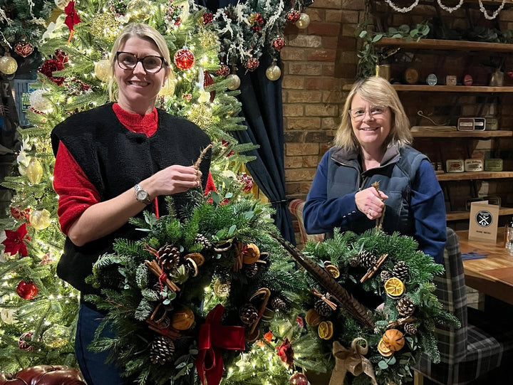 Two smiling women holding freshly assembled Christmas wreaths made with dried orange slices, pinecones, cinnamon sticks, and pheasant feathers.