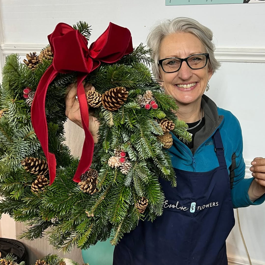 British Flowers Direct owner Helen holding up a complete Christmas wreath made with fresh pine, pinecones, berries, and a bright red velvet ribbon.