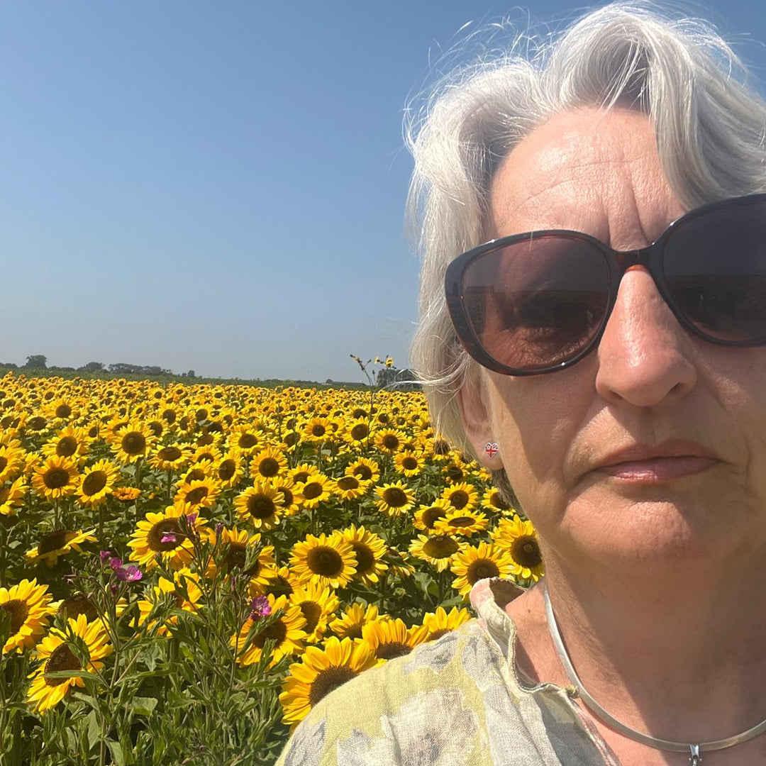 A close-up selfie of Helenwith white hair and dark sunglasses, standing in a vibrant sunflower field under a clear blue sky.