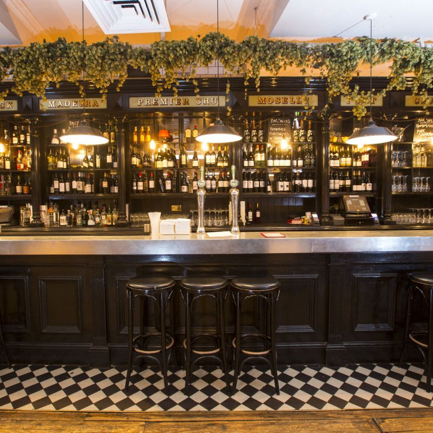 A classic bar interior with a dark wood counter and bar stools, shelves of liquor bottles, hanging pendant lights, and a black-and-white checkered floor. Decorative greenery lines the top of the shelving.