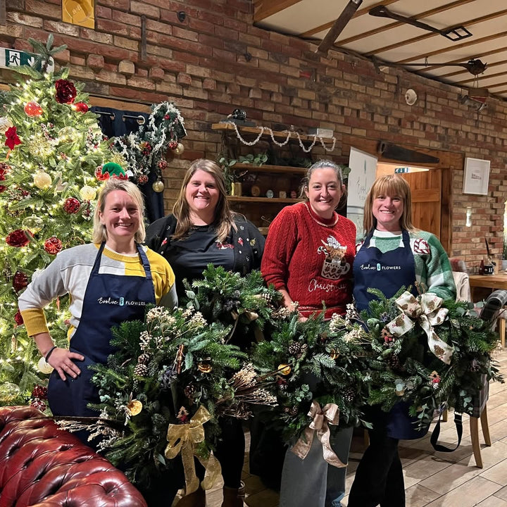 Four women proudly standing together holding lush evergreen Christmas wreaths decorated with pinecones, dried fruits, and decorative ribbons.