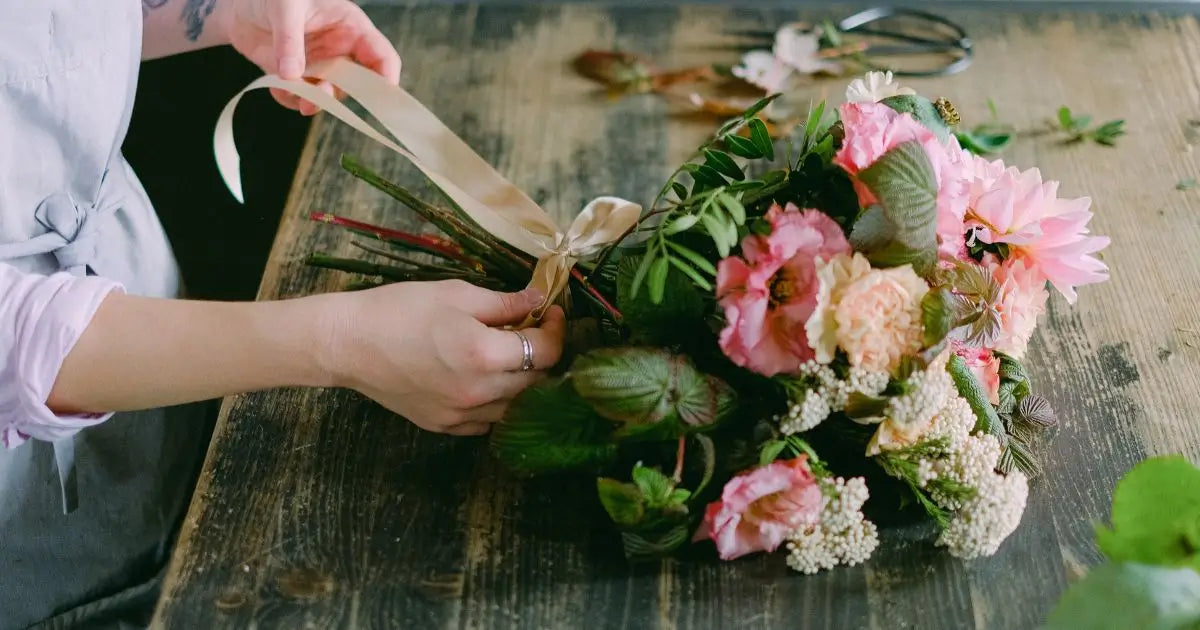 A close-up of a person's hands wrapping a bouquet of pink and white flowers with green leaves in a rustic wooden setting.