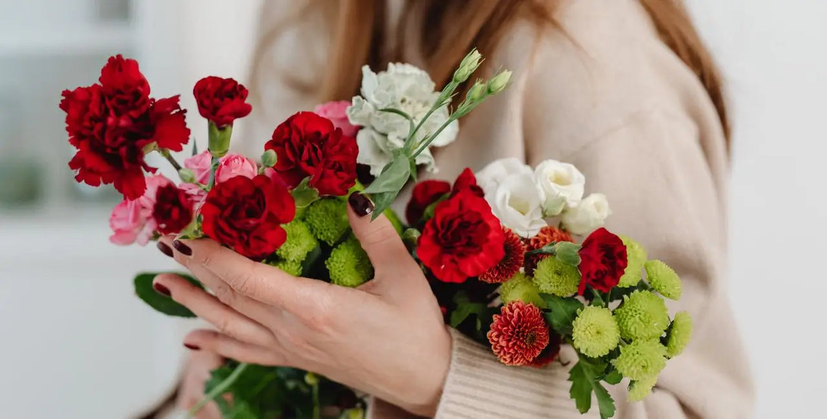 A close-up of a person holding a vibrant bouquet of fresh flowers, including red carnations, white roses, and green chrysanthemums, wearing a beige knit sweater.