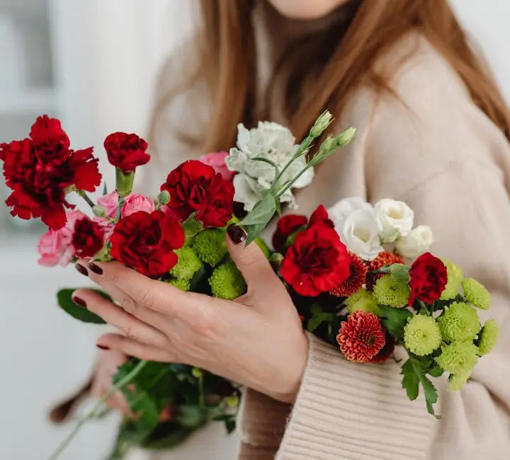 A close-up of a person holding a vibrant bouquet of fresh flowers, including red carnations, pink roses, white lilies, and green chrysanthemums, wearing a beige knit sweater.