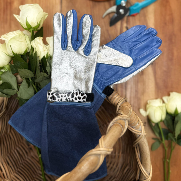 Spinsterz Jayde royal blue and silver leather gardening gauntlets displayed on a rustic table with white roses, blue secateurs, and a potted plant.