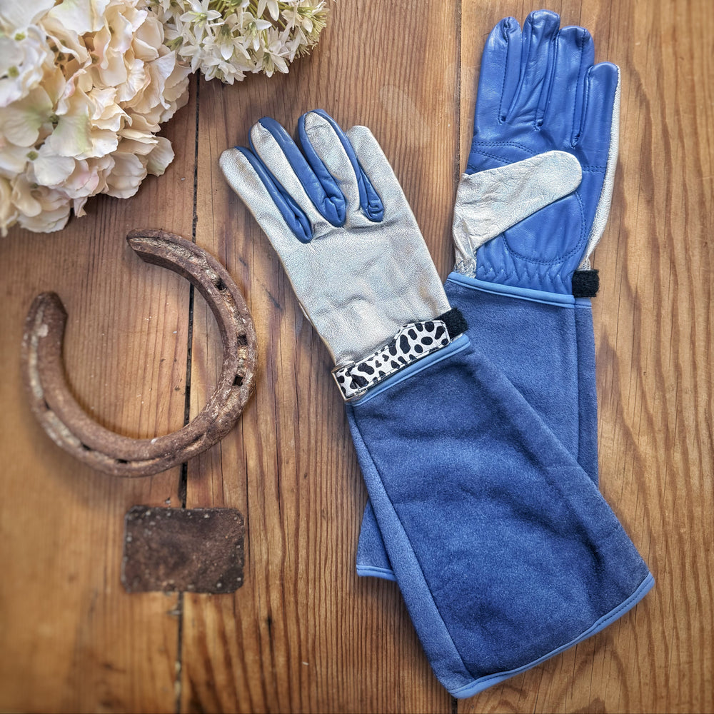 Flat lay arrangement of Spinsterz Jayde royal blue suede gauntlet gloves with silver leather palm and leopard-print trim on a wooden surface next to dried hydrangea flowers.