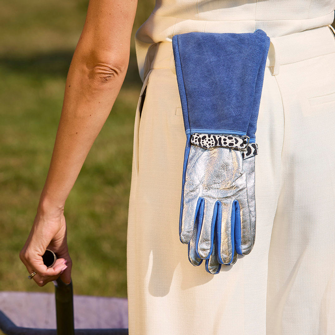 Rear view of a woman outdoors in a white outfit with Spinsterz royal blue, silver, and leopard-print Jayde full-length gauntlet gloves tucked into her back waistband.