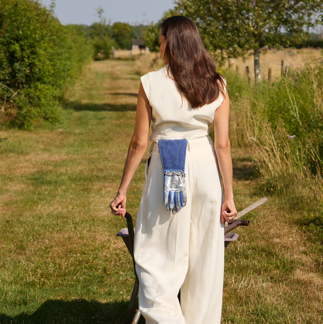 Full-length shot of a woman in a white jumpsuit carrying a garden tool and holding Spinsterz long royal blue gauntlet gloves while walking down a countryside garden path.