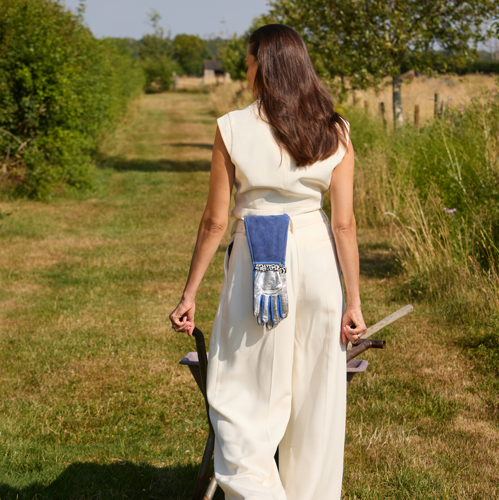 Full-length shot of a woman in a white jumpsuit carrying a garden tool and holding Spinsterz long royal blue gauntlet gloves while walking down a countryside garden path.