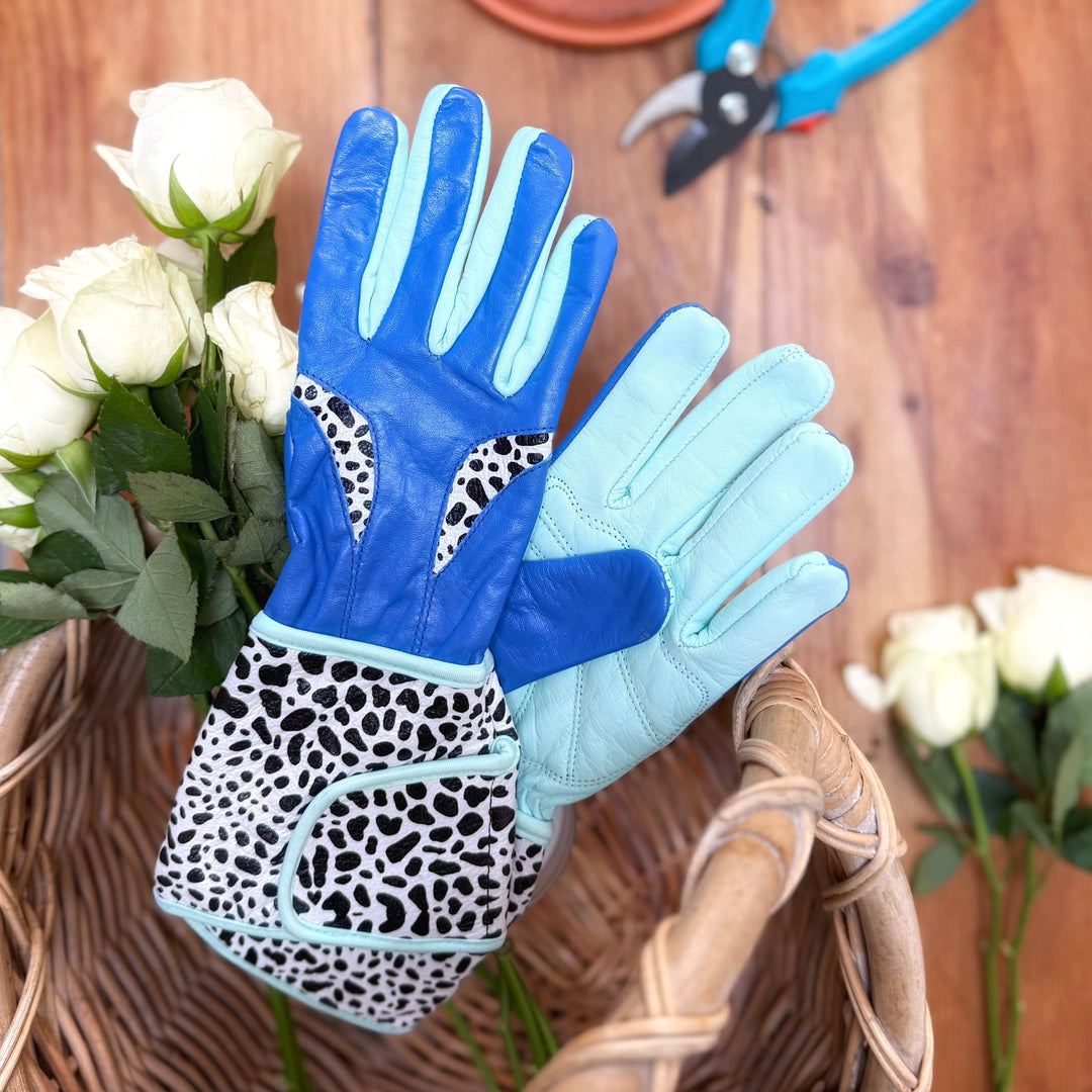 Spinsterz blue and leopard-print leather gauntlet gloves displayed on a rustic wooden table alongside white garden roses, secateurs, and a flower basket.