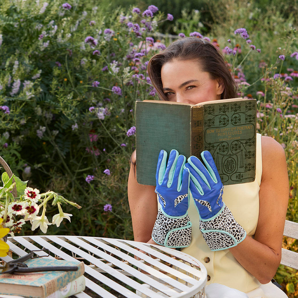A woman sitting outdoors at a garden table holding an open vintage book over her face, wearing Spinsterz sky blue and turquoise MidiGaunt leather gardening gloves with leopard-print cuffs.