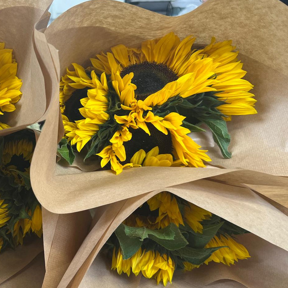 Three small sunflower bouquets wrapped in brown kraft paper, with bright yellow petals, dark brown centers, and green leaves visible.