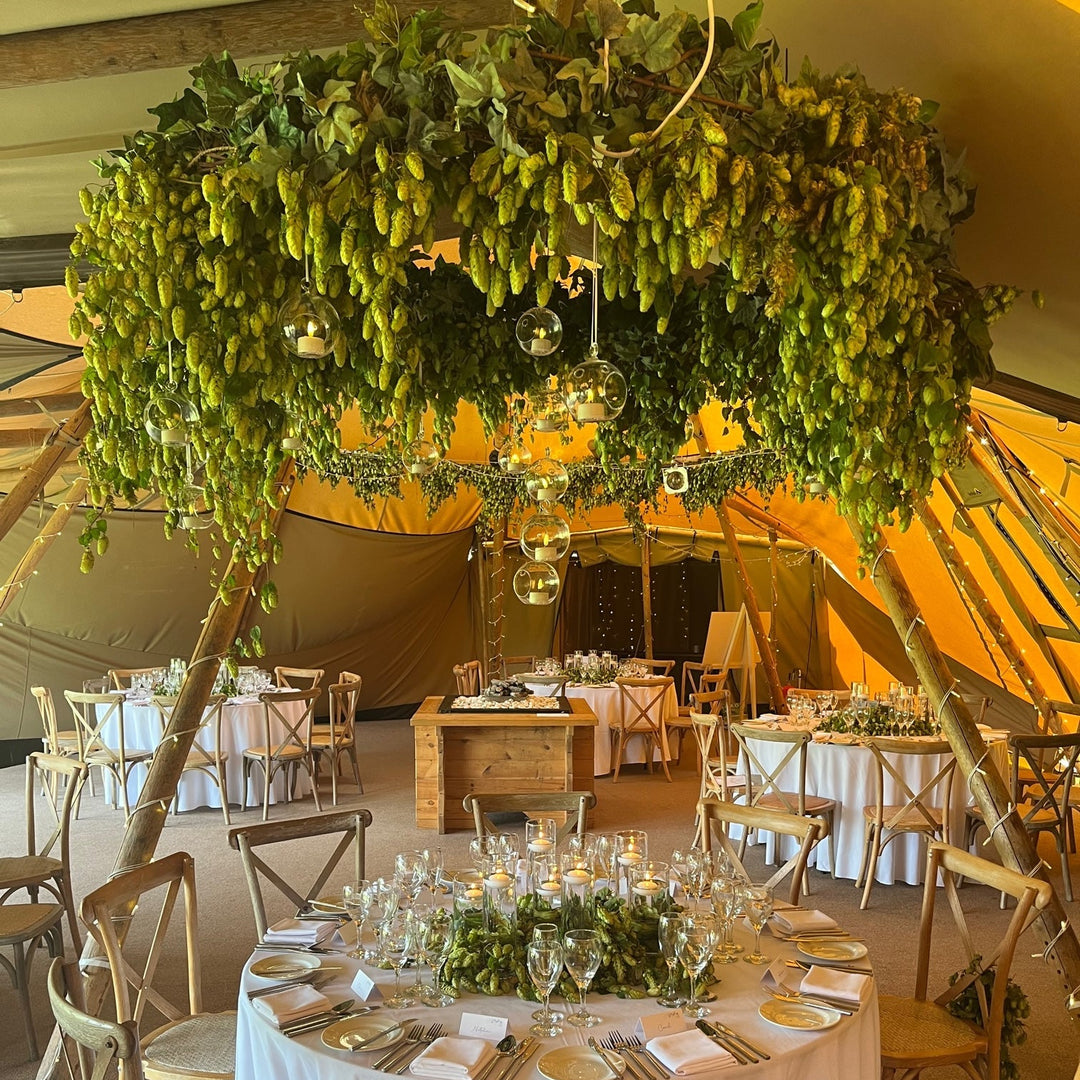 A grand floral chandelier crafted from lush green fresh hop bines hanging above elegant wedding reception tables set inside a wooden tipi tent.