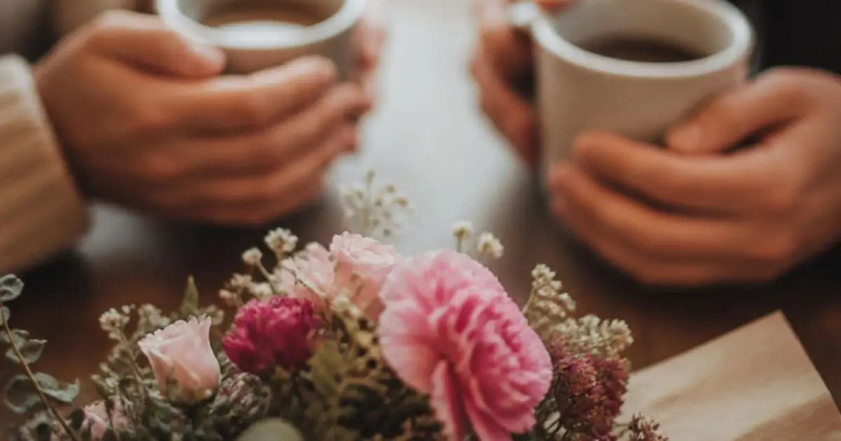 Two hands holding white ceramic mugs filled with coffee at a wooden table, with a pink and purple flower arrangement in the foreground.