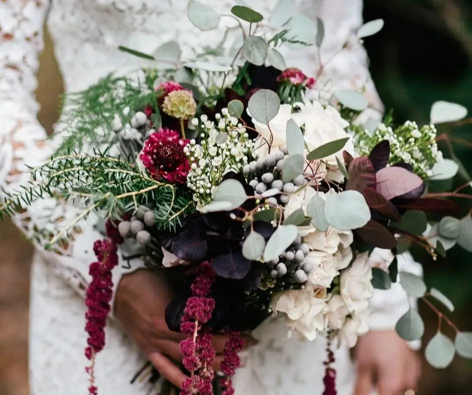 A close-up of a hand holding a lush bridal bouquet with white flowers, green eucalyptus, burgundy accents, and dark foliage, worn by a person in a white lace dress.