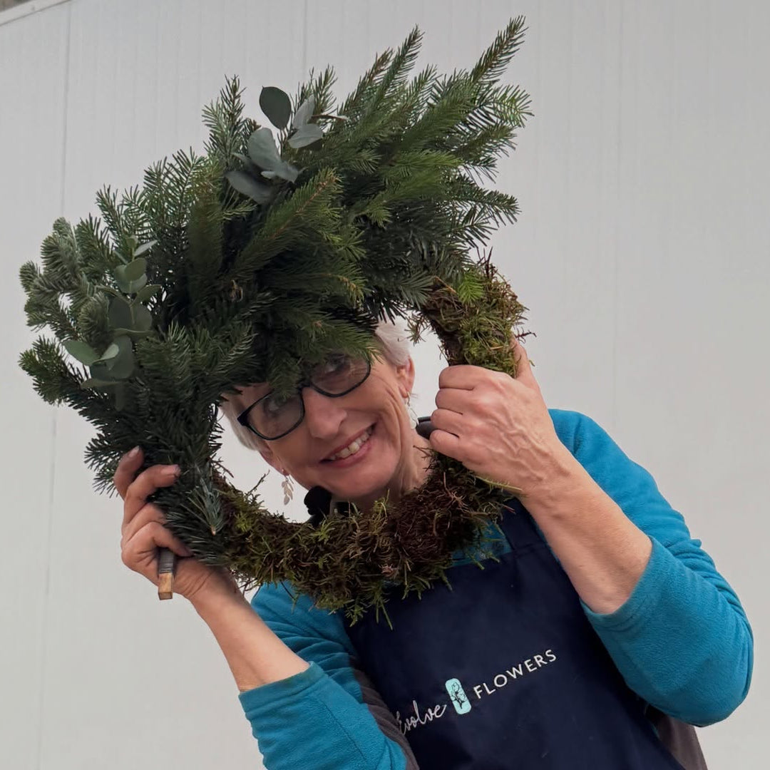 orkshop leader Helen peeking playfully through a handmade moss and pine Christmas wreath base during a British Flowers Direct workshop.