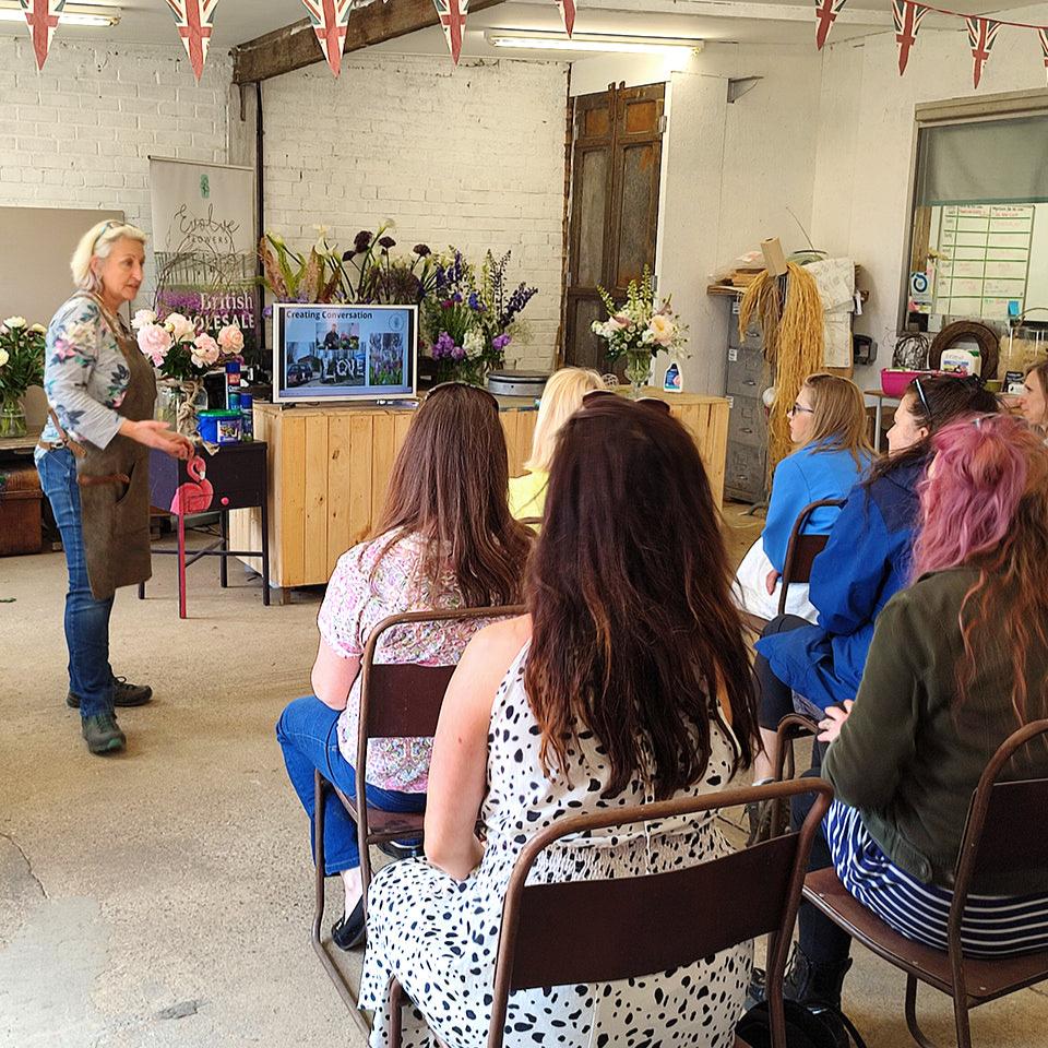A floristry workshop in progress with a group of people seated and listening to a tutor demonstrate how to arrange flowers.