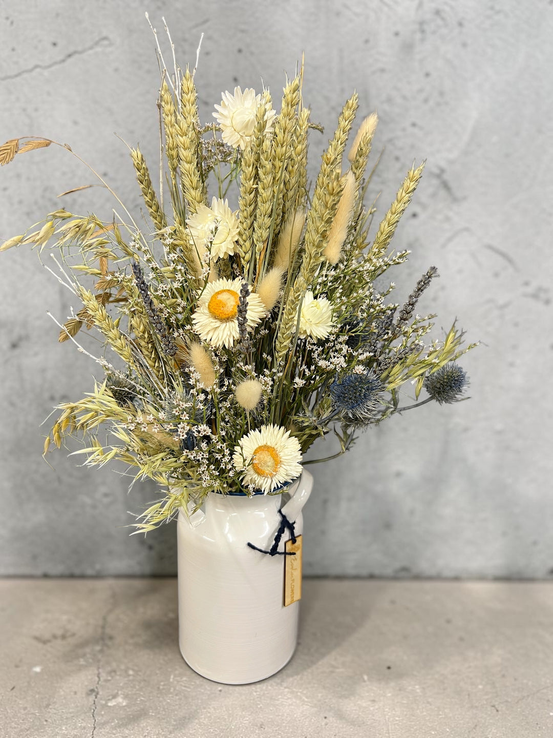 A rustic dried flower arrangement featuring wheat stalks, daisies, and blue thistles in a white ceramic pitcher vase, set against a textured gray concrete wall.