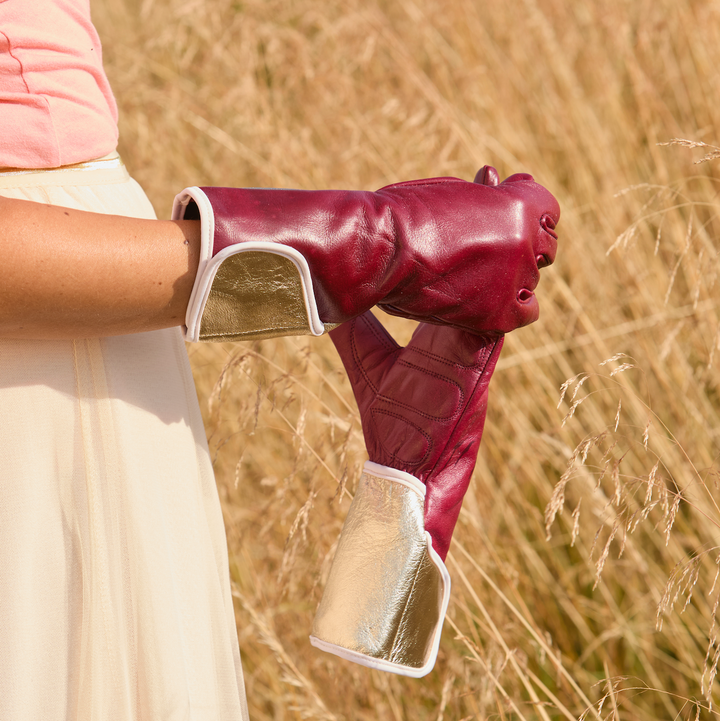Woman wearing the Carol MidiGaunt gardening glove in claret leather with metallic gold cuff, holding the matching pair in a field.
