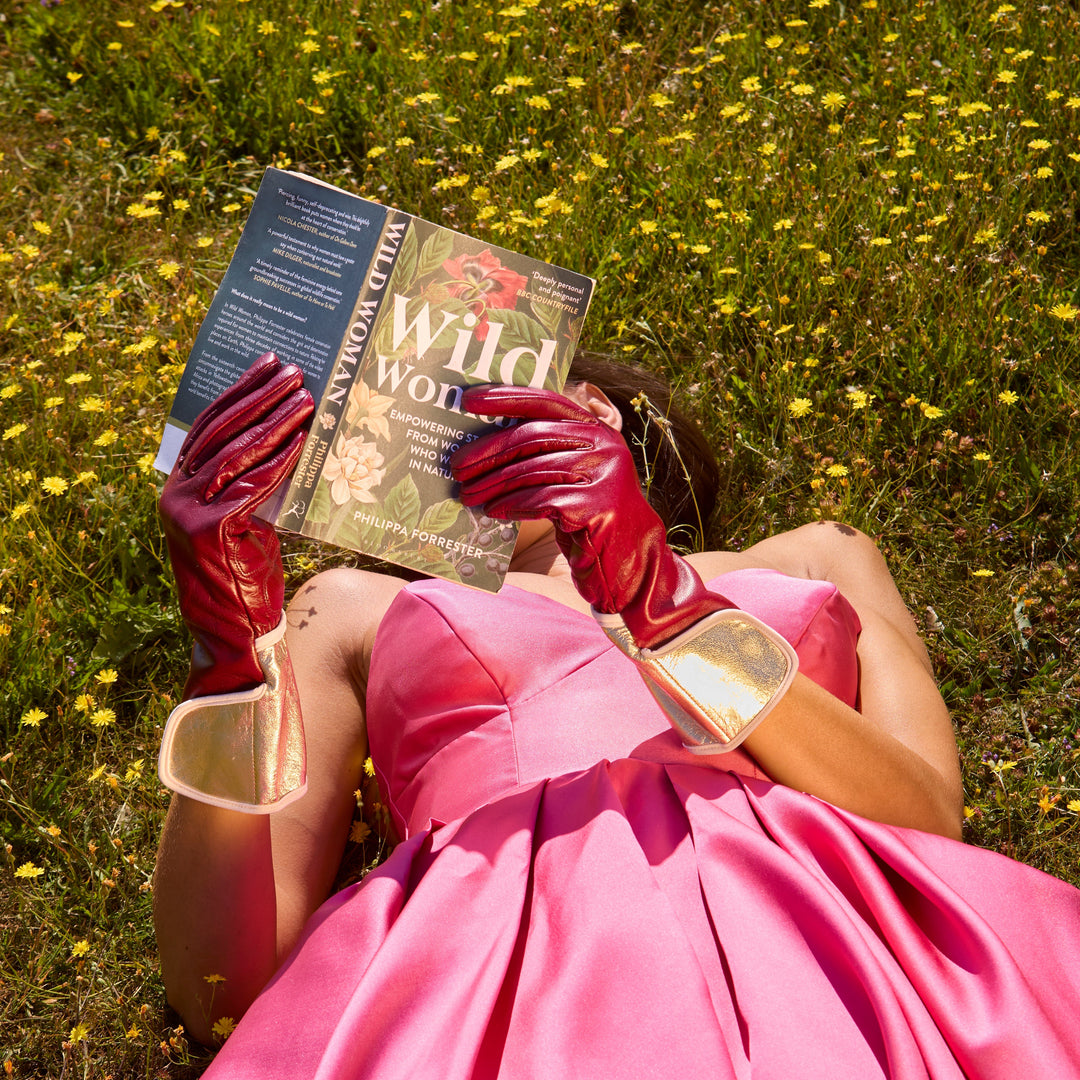 Woman lying in a meadow wearing claret leather Carol gardening gloves and holding a book.
