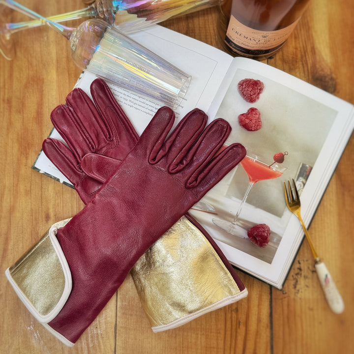 Pair of claret and gold Carol MidiGaunt gardening gloves styled on a wooden table alongside an open magazine and champagne glasses.