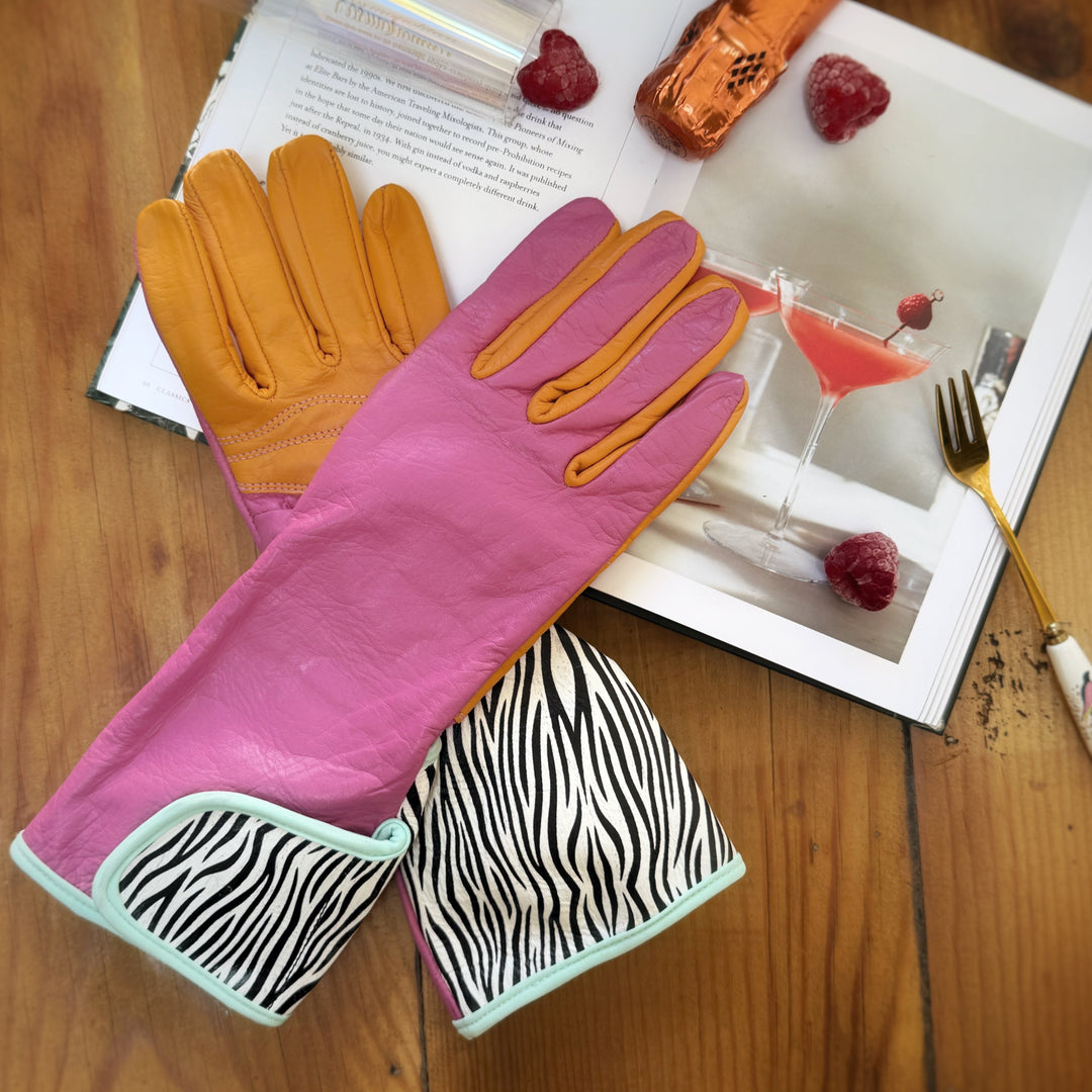 Pair of pink and orange Hannah MidiGaunt gloves with zebra cuffs styled on a wooden table with champagne and fresh raspberries